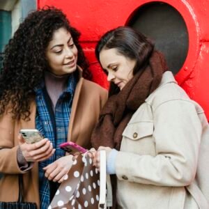 Positive young diverse female best friends in stylish warm clothes standing on city street near red door and browsing mobile phones