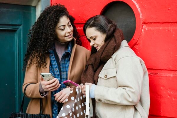 Positive young diverse female best friends in stylish warm clothes standing on city street near red door and browsing mobile phones